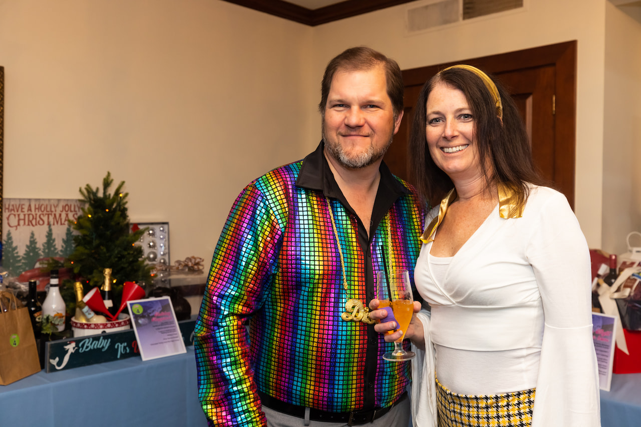 A man and a woman posing together at a festive event, the man wearing a colorful shirt and holding a wine glass, with christmas decorations in the background.