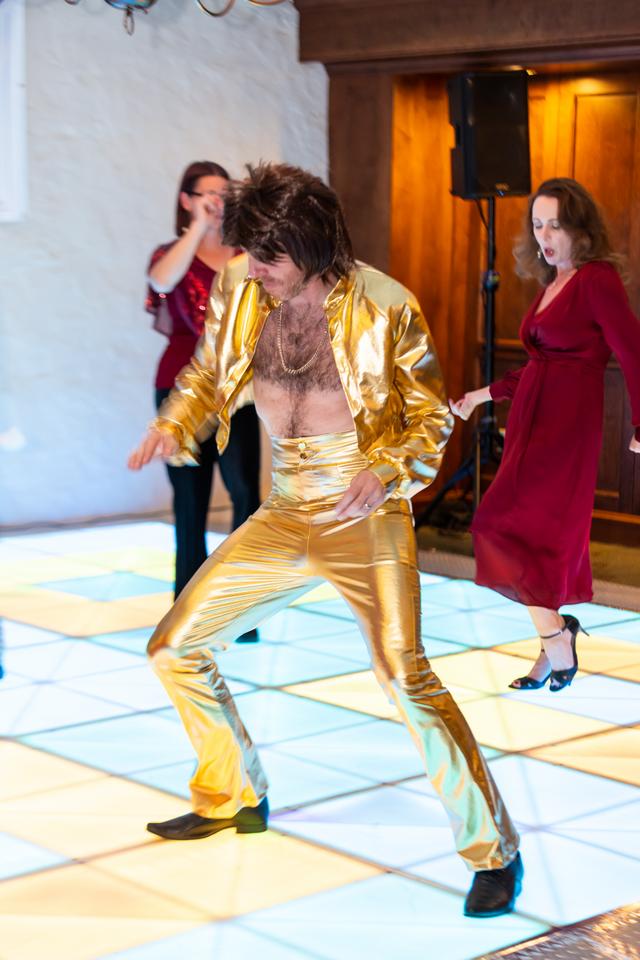Man in a gold suit dancing on a lit dance floor with two women in the background.