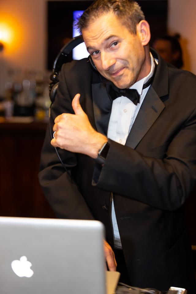 Dj in a black suit giving a thumbs-up behind his laptop setup at an event.