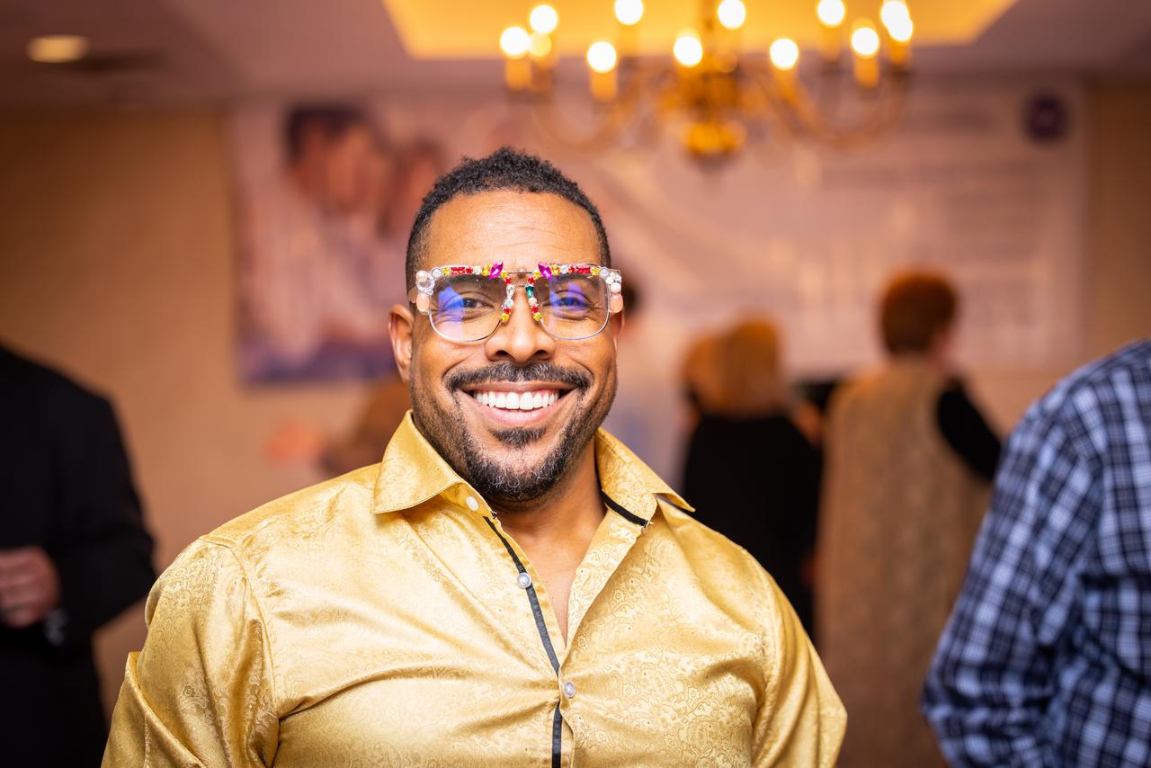 A smiling man wearing a gold shirt and festive glasses at a social event.